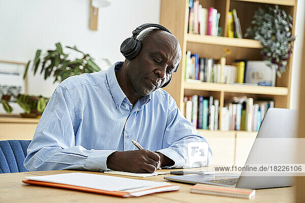 Mature African-American man taking notes at remote meeting wearing headphones while working at home with laptop computer