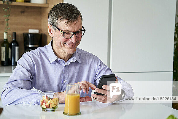 Middle-aged professional man smiling and checking messages on phone while having breakfast on kitchen counter at home