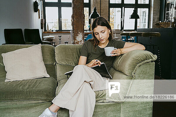 Junger Unternehmer liest ein Buch und trinkt einen Kaffee  während er auf dem Sofa im Studio sitzt
