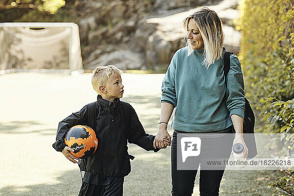 Mutter hält die Hand ihres Sohnes  der einen Fußball trägt  während er auf dem Boden läuft