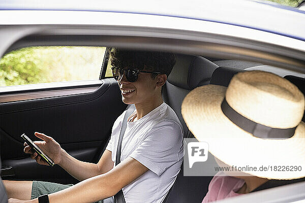 Smiling teenage boy with smart phone wearing sunglasses sitting by brother in car