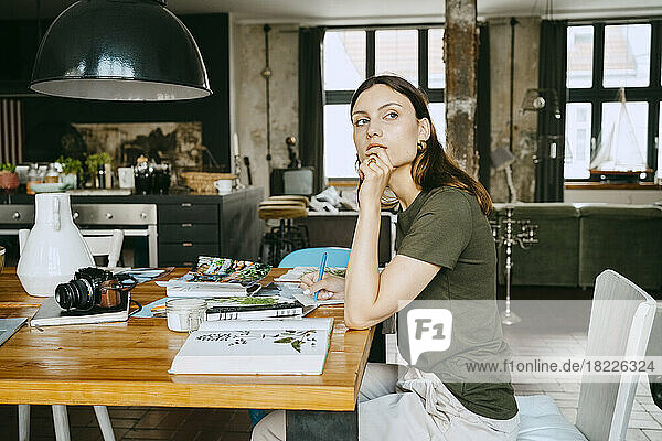 Female food stylist with hand on chin looking away while sitting at table in studio