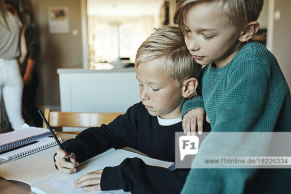 Junge mit Hand auf der Schulter seines Bruders  der zu Hause Hausaufgaben in einem Buch macht
