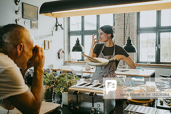 Männlicher Fotograf fotografiert eine Köchin  die in einer Studioküche eine Pfanne hält und gestikuliert