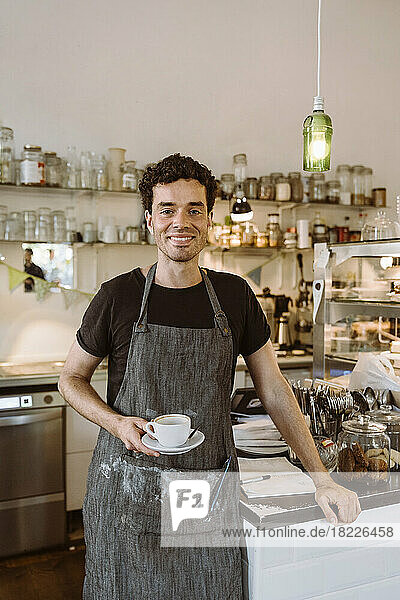 Smiling male owner holding coffee cup standing in cafe