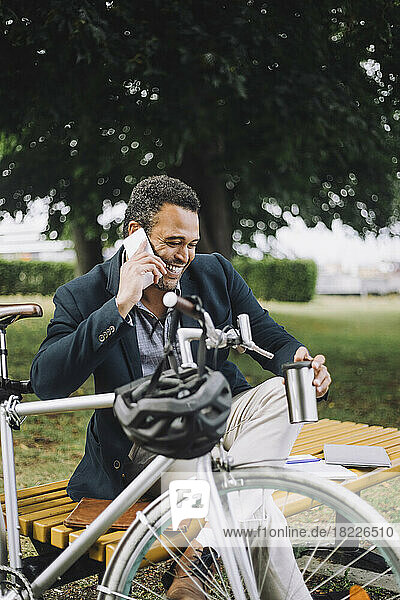 Happy businessman talking on mobile phone while sitting by bicycle at park