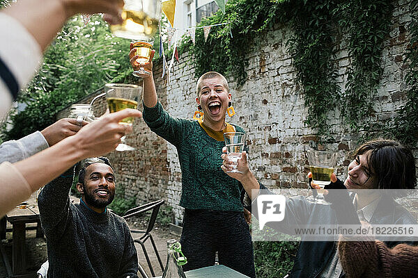 Fröhliche Frau mit offenem Mund stößt mit Freunden während einer Dinnerparty im Hinterhof auf Getränke an