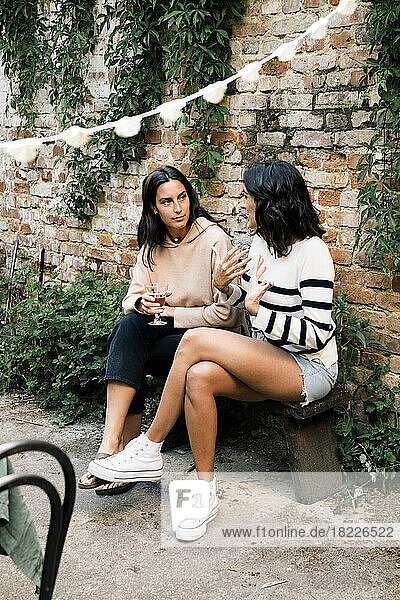 Female friends talking to each other while sitting on bench near brick wall at garden party
