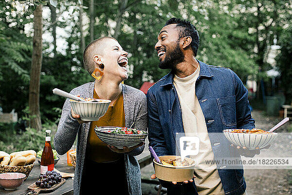 Multiracial male and female friends laughing and looking at each other while holding food bowls