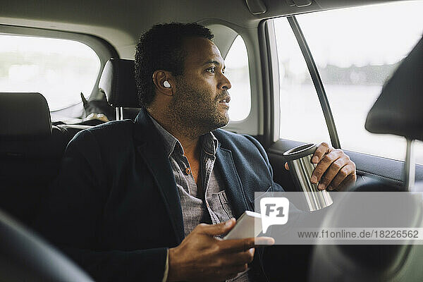 Männlicher Unternehmer mit Mobiltelefon und isoliertem Getränkebehälter im Auto sitzend
