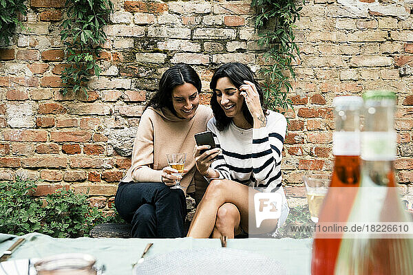 Lächelnde Freundinnen  die sich ein Smartphone teilen  sitzen auf einer Bank an einer Backsteinmauer bei einer Gartenparty