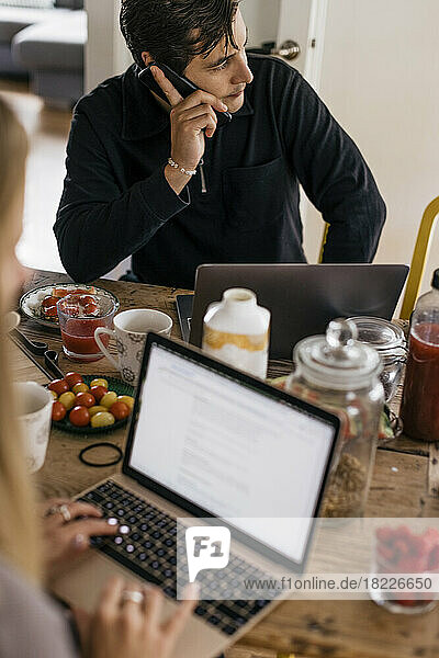 Männlicher Freiberufler  der mit seinem Smartphone telefoniert  während er zu Hause auf dem Tisch sitzt und frühstückt