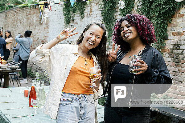 Porträt von glücklichen Freundinnen mit Getränken und Friedenszeichen während einer Dinnerparty im Hinterhof