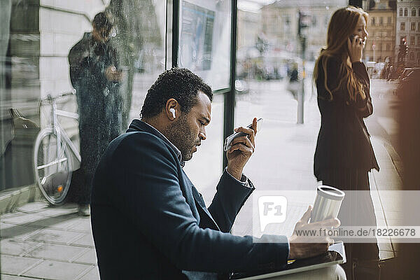 Seitenansicht eines männlichen Berufstätigen  der eine Voicemail sendet  während er an einer Bushaltestelle gegenüber einer Geschäftsfrau sitzt  die mit einem Mobiltelefon spricht