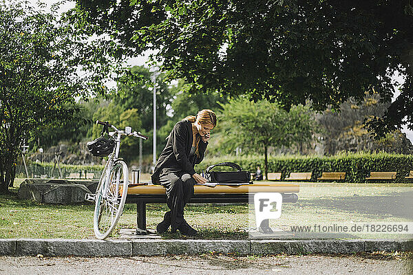 Full length of businesswoman talking on smart phone while sitting by bicycle at park