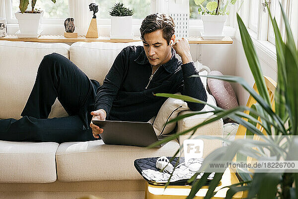 Young male freelancer watching laptop while lying on sofa at home
