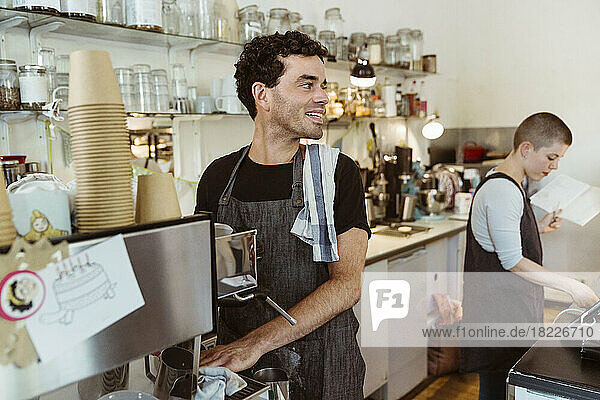 Lächelnder Besitzer mit Schürze  der wegschaut  während er in der Nähe der Kaffeemaschine im Café steht