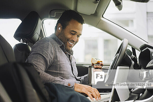 Lächelnder Geschäftsmann mit isoliertem Getränkebehälter  der einen Laptop benutzt  während er im Auto sitzt