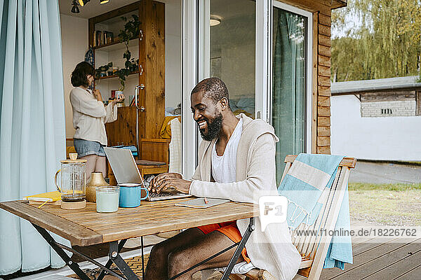 Glücklicher reifer Mann  der einen Laptop benutzt  während er am Tisch auf der Veranda vor dem Haus sitzt