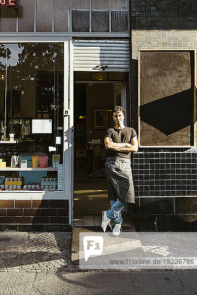 Male owner with arms crossed standing outside cafe on sunny day
