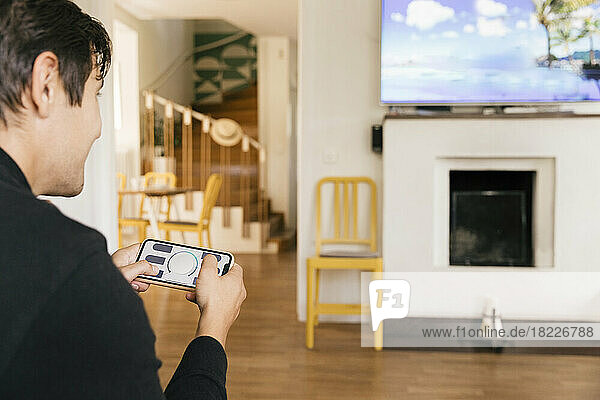 Mann  der eine Smart-Home-Anwendung auf seinem Mobiltelefon benutzt  während er im heimischen Wohnzimmer fernsieht