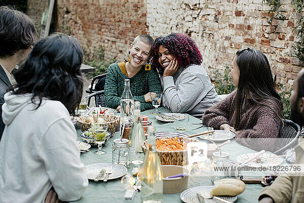 Friends talking to multiracial women sitting at dining table during dinner party in back yard