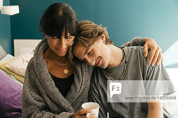 Smiling mother with arm around son leaning on shoulder sitting on bed at home