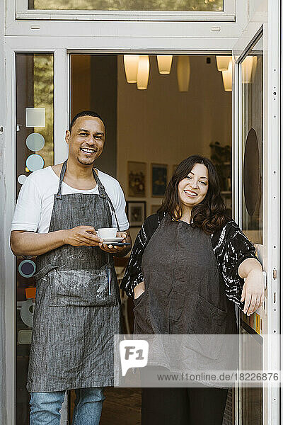 Portrait of happy multiracial male and female cafe owners standing at doorway