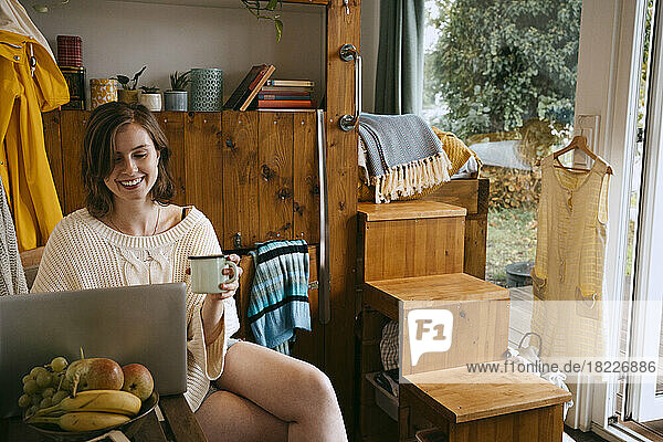 Lächelnde junge Frau mit Laptop und Kaffeetasse  während sie zu Hause sitzt