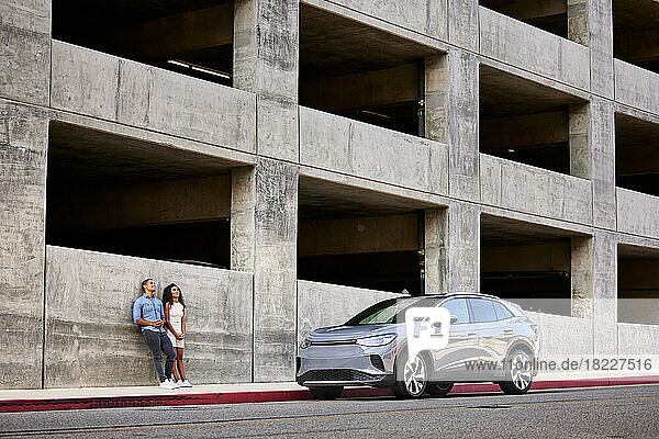 Couple leaning on building wall by electric car parked at roadside