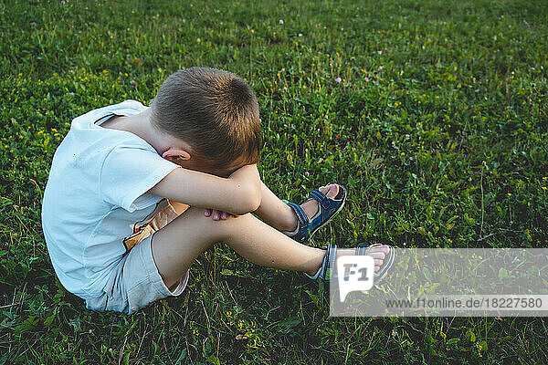 Boy is sitting on the grass and covering face with his hands