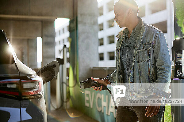 Man in denim jacket holding electric plug by car at charging station