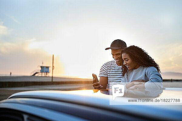 Smiling heterosexual couple using smart phone by car during sunset