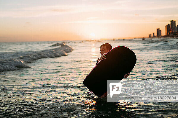 Young boy holding board in ocean waves on beach at sunset