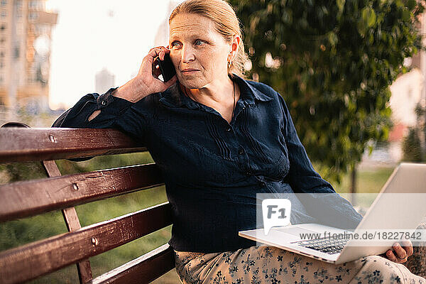 Mature business woman talking on mobile phone holding laptop in city