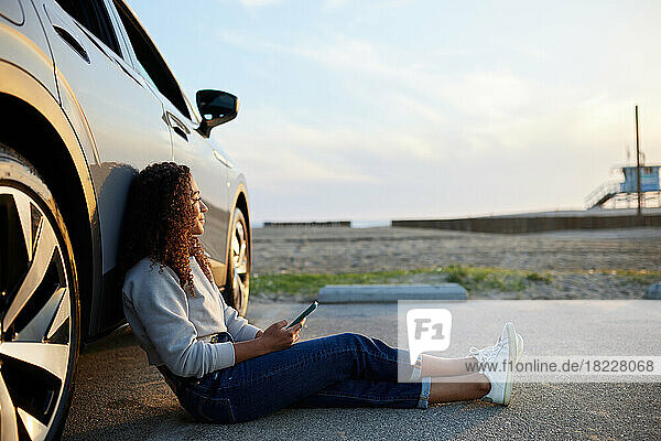Young woman with smart phone leaning on electric car at parking lot