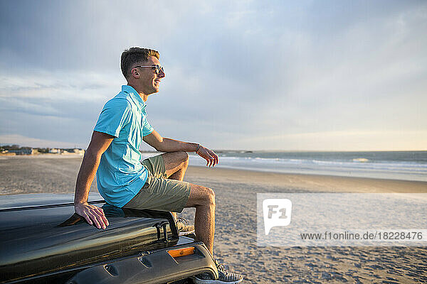 Young Man with sunglasses on hood of car at sunrise at Beach