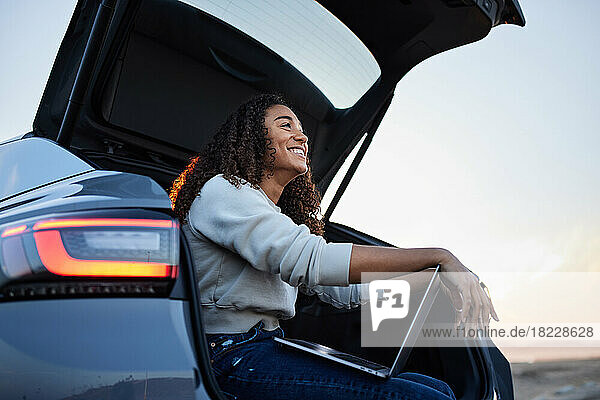 Happy woman with laptop sitting in car trunk