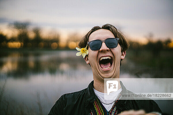 Teenage boy laughs with sunglasses on by lake at sunset