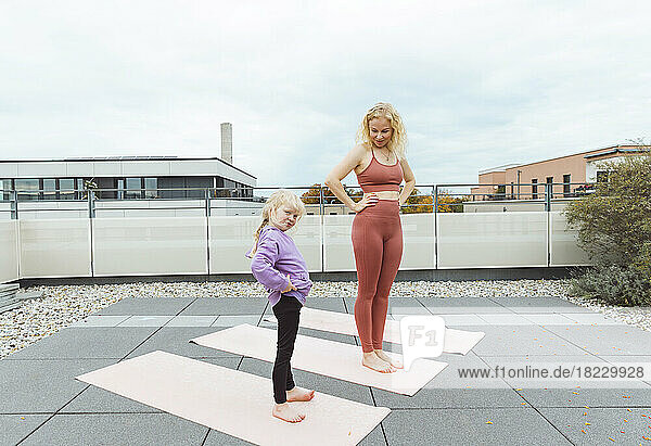 Glückliche Mutter und Tochter mit den Händen auf den Hüften  die auf dem Dach Yoga praktizieren