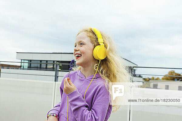 Fröhliches Mädchen hält eine kleine Blume in der Hand und hört Musik über Kopfhörer auf dem Dach