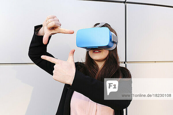 Young woman wearing virtual reality simulator gesturing in front of wall