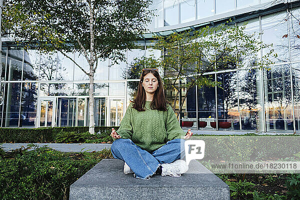 Young woman with eyes closed meditating on concrete seat in front of glass building