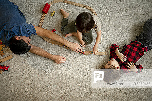 Junge schaut Vater und Bruder an  die mit einer hölzernen Spielzeugeisenbahn auf dem Boden spielen
