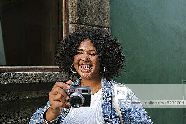 Fröhliche junge Frau mit lockigem Haar hält eine analoge Kamera vor der Wand