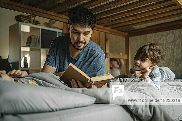 Father reads book to his sons on bed at home