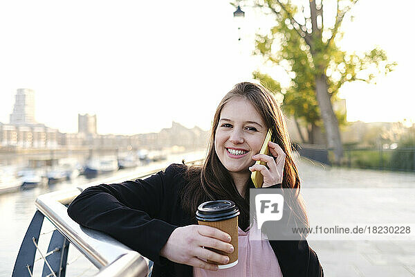 Happy young woman holding disposable cup talking on phone by railing