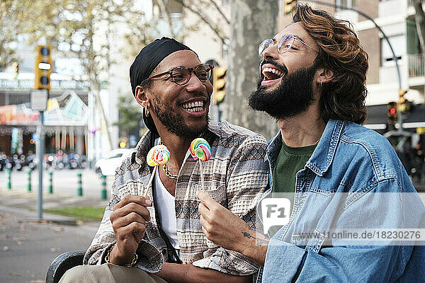 Happy gay couple enjoying candy sitting on bench
