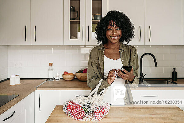 Smiling woman holding smart phone with mesh bag on kitchen island at home