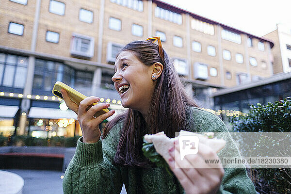 Happy woman holding sandwich talking through speaker of mobile phone in front of building